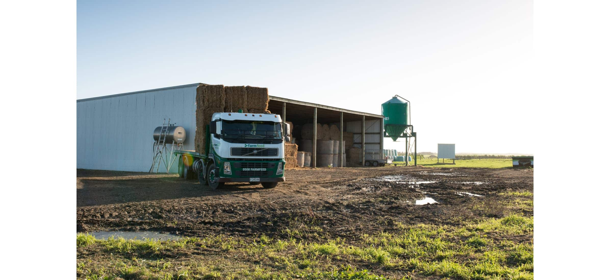 Long truck filled with hay bales parked next to a farm shed.